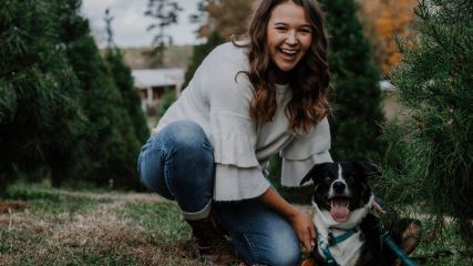 A photograph of Lauren crouching next to her dog among pine trees