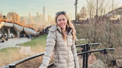 A photo of Amy wearing a cream-colored jacket outdoors in a park, with a city skyline in the background