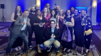 A group of 14 BrandExtract employees standing in a group in an event center, holding up their awards form the 2023 Crystal Awards.