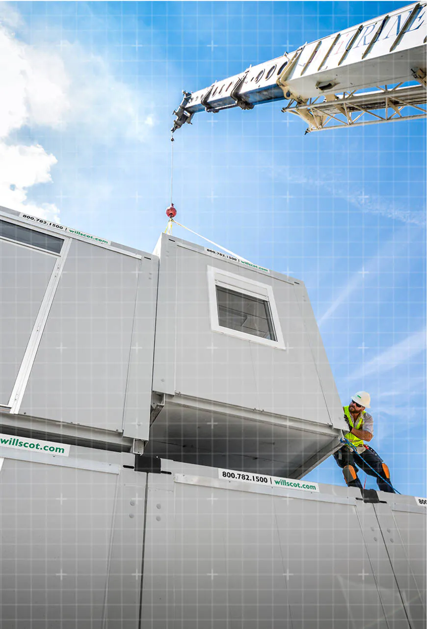 A WillScot modular space being moved into place by a crane with a man guiding it down into position